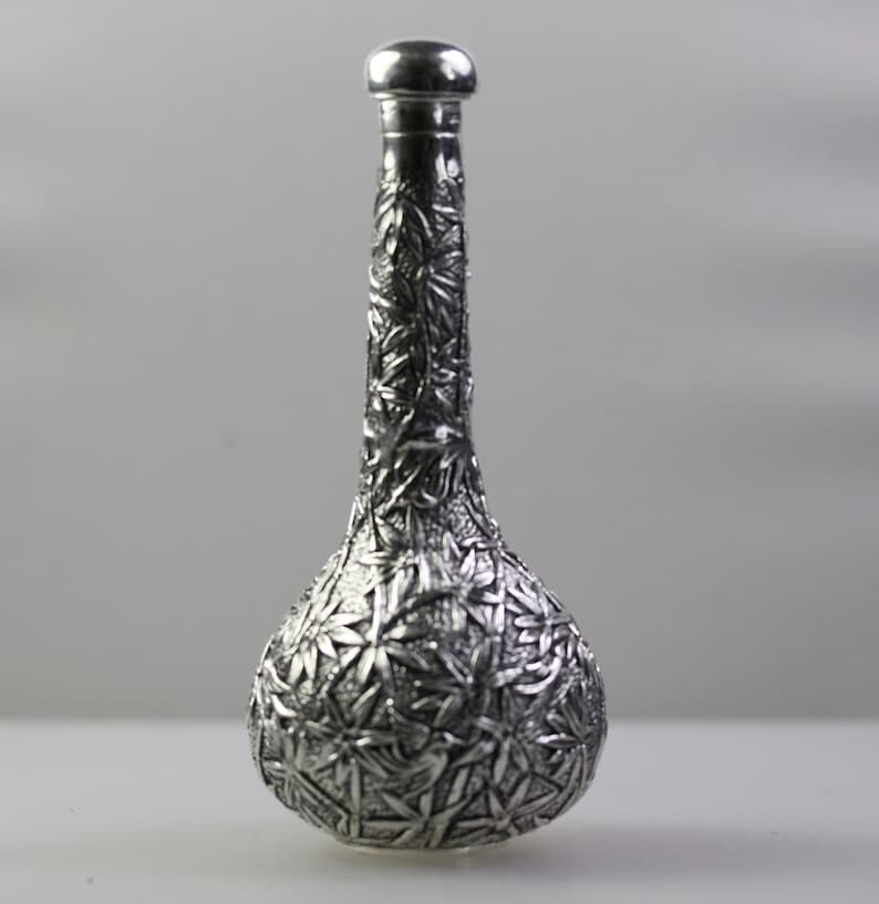 May include: A silver decorative bottle with an intricate floral design. The bottle has a round base and a long, slender neck.