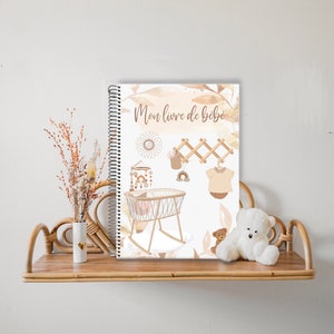 Peut inclure: Un livre de bébé beige et marron avec le titre "Mon livre de bébé" et des illustrations d'un berceau, d'un mobile, d'un porte-vêtements et d'une tenue de bébé.