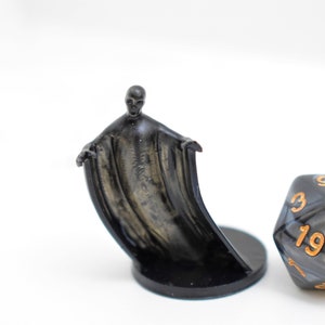 May include: A black and silver miniature figurine of a ghost with a flowing cape, standing on a black base. A black and silver d20 dice with gold numbers is next to the figurine.