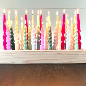 May include: A collection of lit, colorful twist candles in a wooden holder. The candles are in shades of pink, green, yellow, and white. The wooden holder is rectangular and made of light-colored wood. The candles are arranged closely together.