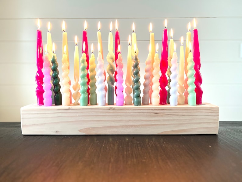 May include: A collection of lit, colorful twist candles in a wooden holder. The candles are in shades of pink, green, yellow, and white. The wooden holder is rectangular and made of light-colored wood. The candles are arranged closely together.