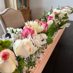 May include: A wooden centerpiece with a series of small glass vials, each holding a fresh flower. The arrangement includes white roses, pink roses, and white chrysanthemums, creating a vibrant floral display.