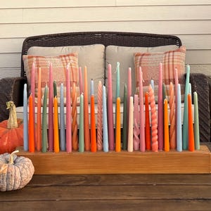 May include: A wooden candle holder with a variety of colorful taper candles. The candles are in shades of pink, orange, green, and blue. The holder is on a wooden table with pumpkins in the foreground.
