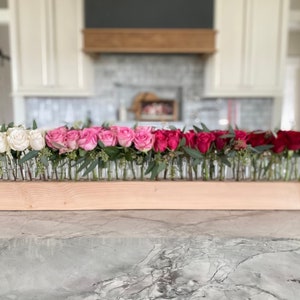 May include: A floral arrangement featuring white, pink, and red roses in individual glass vials. The vials are arranged in a row on a light-colored wooden base, with greenery accents. The arrangement is a decorative centerpiece.