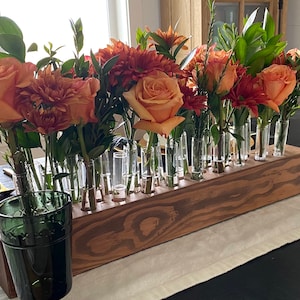 May include: A wooden flower vase with multiple glass tubes holding an arrangement of orange and red flowers and greenery. The vase is rectangular and sits on a table with a white runner. A green glass is in the foreground.