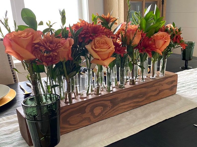 May include: A wooden flower vase with multiple glass tubes holding an arrangement of orange and red flowers and greenery. The vase is rectangular and sits on a table with a white runner. A green glass is in the foreground.