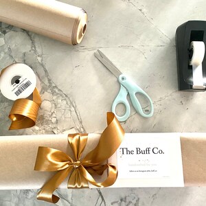 May include: A gift-wrapping scene with a roll of brown paper, gold ribbon, and a pair of light blue scissors. A package is wrapped in brown paper and tied with a gold satin bow. A label reads "The Buff Co."