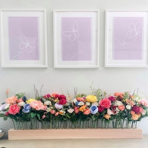 May include: Three framed butterfly art prints above a floral arrangement. The prints feature white line drawings of butterflies on a lavender background. The floral arrangement has a variety of colorful flowers in individual glass tubes set in a wooden base.