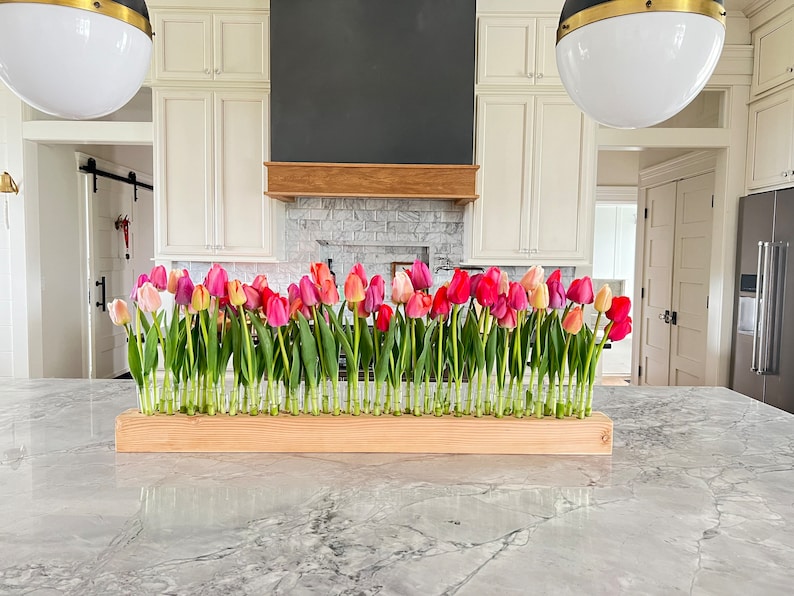 May include: A floral arrangement of colorful tulips in a wooden trough. The tulips, in shades of pink, red, and yellow, are displayed in individual glass vials. The arrangement sits on a marble countertop in a kitchen setting.