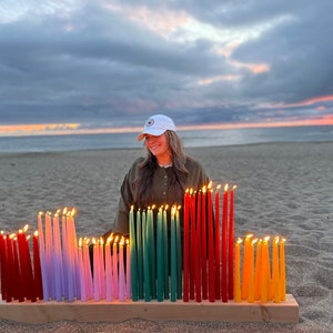 May include: A wooden candle holder with multiple lit taper candles in various colors, including red, purple, pink, green, orange, and yellow. The candles are arranged in rows on a wooden base, creating a colorful display. The scene is set on a sandy beach at sunset.
