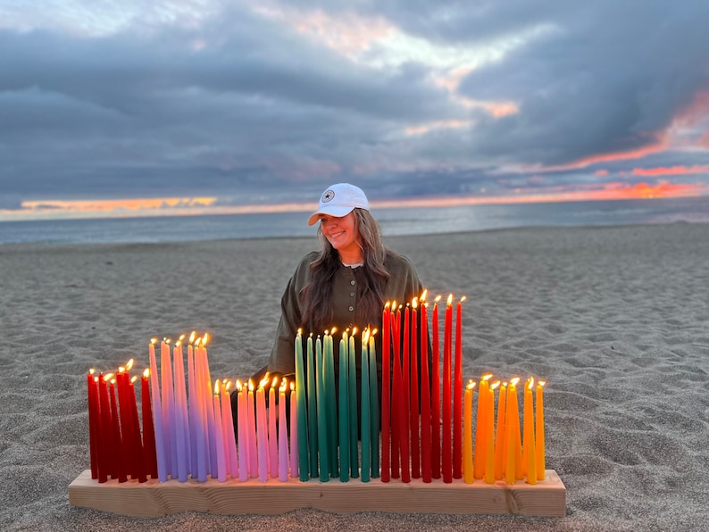 May include: A wooden candle holder with multiple lit taper candles in various colors, including red, purple, pink, green, orange, and yellow. The candles are arranged in rows on a wooden base, creating a colorful display. The scene is set on a sandy beach at sunset.
