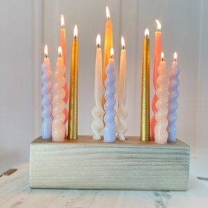 May include: A collection of lit candles in various colors, including gold, pink, and lavender, arranged in a wooden holder. The candles have different shapes, with some being twisted and others straight. The candles are on a white marble table.