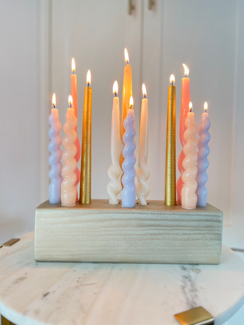 May include: A collection of lit candles in various colors, including gold, pink, and lavender, arranged in a wooden holder. The candles have different shapes, with some being twisted and others straight. The candles are on a white marble table.
