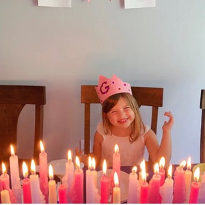 May include: A young person wearing a pink crown with the letter "G" smiles brightly at a table. Lit candles in shades of pink and white are arranged in front of the person. The scene suggests a birthday celebration.
