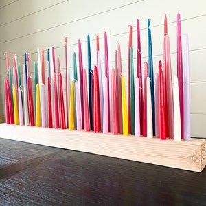 May include: A wooden candle holder with a variety of colorful taper candles. The candles are in shades of red, pink, yellow, green, blue, and purple. The holder is made of light-colored wood.