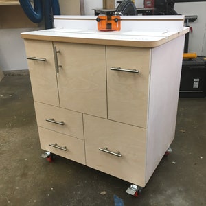 May include: A light-coloured wooden router table cabinet featuring a flat top surface with an orange fence guard, two upper doors, and three lower drawers. The unit is equipped with silver metal handles and is mounted on four locking caster wheels.