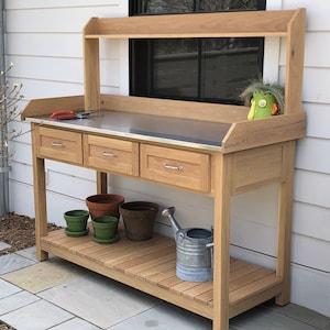 May include: A wooden potting bench featuring a stainless steel countertop, three drawers with silver-toned handles, and an upper shelf. The lower slatted shelf holds two green pots, two brown terracotta pots, and a galvanized metal watering can.