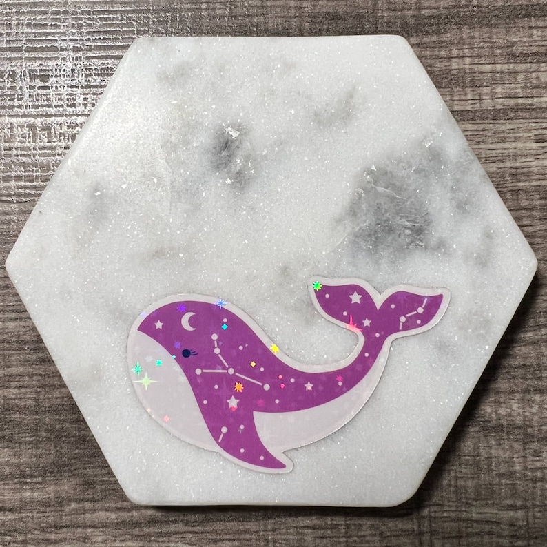 May include: A white hexagonal coaster with a purple whale sticker. The whale has a constellation design with stars and a crescent moon. The coaster has a glittery surface.