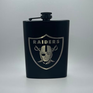 May include: Black flask with a silver Raiders logo featuring a helmet, crossed swords, and the word "RAIDERS".
