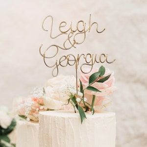 May include: Two white wedding cakes decorated with pink and white flowers and greenery. The top cake features a gold wire topper that reads "Leigh & Georgia".