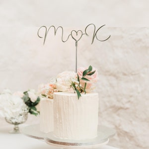 May include: Two-tiered wedding cake with white textured frosting, decorated with pink roses and greenery. A silver wire cake topper spells "m♡k". The cake is on a wooden stand with a silver base. The cake is a centerpiece.
