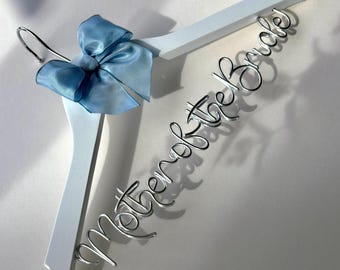 May include: A white wooden clothes hanger featuring a silver wire script that reads Mother of the Bride. The hanger is decorated with a light blue satin ribbon bow tied at the top near the chrome metal hook, creating a decorative display piece.