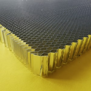 Puede incluir: Primer plano de una estructura metálica de panal de abeja. La capa superior es plateada y los lados dorados. El patrón hexagonal crea un diseño geométrico. El material parece rígido y duradero, adecuado para diversas aplicaciones.