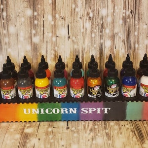 May include: A display of 15 bottles of Unicorn Spit paint in various colors, including purple, brown, gray, yellow, teal, red, gold, green, blue, and white. The bottles are arranged in a row on a rainbow-colored wooden shelf with the words "Unicorn Spit" printed in white.
