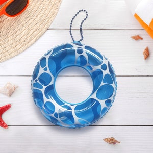May include: A blue and white patterned inflatable swim ring with a chain, next to a straw hat, orange sunglasses, a red starfish, seashells, and an orange and white striped towel. The items are arranged on a white wooden surface.