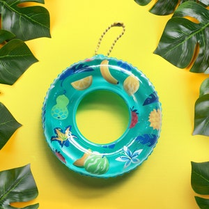 May include: A turquoise inflatable pool ring with fruit illustrations, including a banana, lemon, and watermelon. The ring has a beige handle and is surrounded by large green tropical leaves on a bright yellow background.