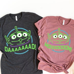 May include: Two t-shirts, one dark gray and one mauve, each featuring a green alien face with three eyes inside a blue circle. The dark gray shirt says "DAAAAAAD!" in green, and the mauve shirt says "MOOOOOM!" in green.