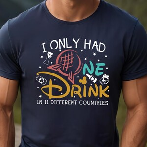 May include: Navy blue t-shirt with the text "I ONLY HAD ONE DRINK IN 11 DIFFERENT COUNTRIES" in white, yellow, and teal. The design includes a stylized globe and beer mugs. The shirt is worn by a person with blonde hair.