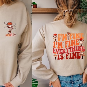 May include: A beige sweatshirt with a graphic of a cartoon character with the word "Anxiety" printed below. The back of the sweatshirt has the text "I'm fine I'm fine Everything is fine" with cartoon characters around the text.