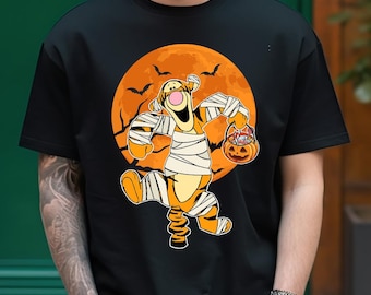 Op de afbeelding: Een zwart T-shirt met een afbeelding van Winnie de Poeh verkleed als een mummie die een jack-o-lantern vasthoudt. De afbeelding is geplaatst tegen een achtergrond van een oranje maan met vleermuizen die eromheen vliegen.