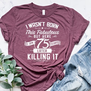 May include: A maroon t-shirt with the text "I wasn't born this fabulous but here I am 75 years later killing it".