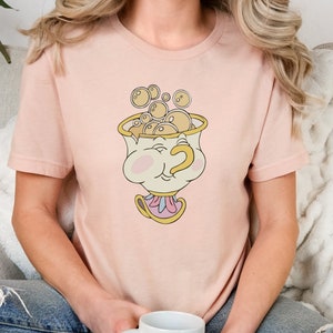 Puede incluir: Una camiseta rosa con una imagen de dibujos animados de una taza de t&eacute; con una cara y burbujas que salen de ella. La taza de t&eacute; tiene un borde dorado y un asa rosa y blanca.