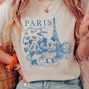 Op de afbeelding: Een licht beige T-shirt met een blauwe afbeelding van Mickey Mouse en Minnie Mouse voor de Eiffeltoren. De tekst "PARIS" is boven de afbeelding gedrukt.