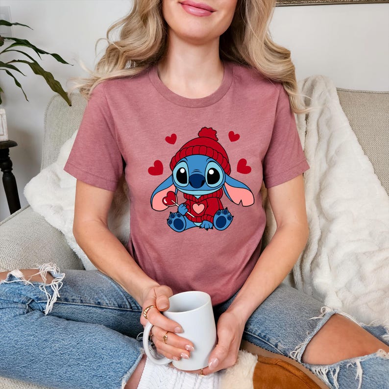 May include: A mauve-colored t-shirt featuring a cartoon character, Stitch, wearing a red knit cap and sweater, holding a heart-shaped lollipop. Red hearts are scattered around the character. The shirt has short sleeves.