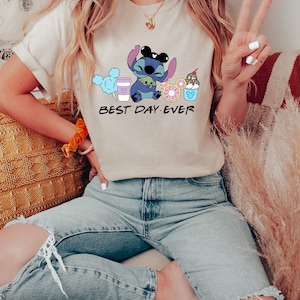 K&ouml;nnte beinhalten: Eine Frau tr&auml;gt ein hellbeiges T-Shirt mit einer blauen Comicfigur, die einen gr&uuml;nen Au&szlig;erirdischen h&auml;lt und von Comicbildern von Essen und Getr&auml;nken umgeben ist. Der Text "BEST DAY EVER" ist unter der Comicfigur gedruckt.