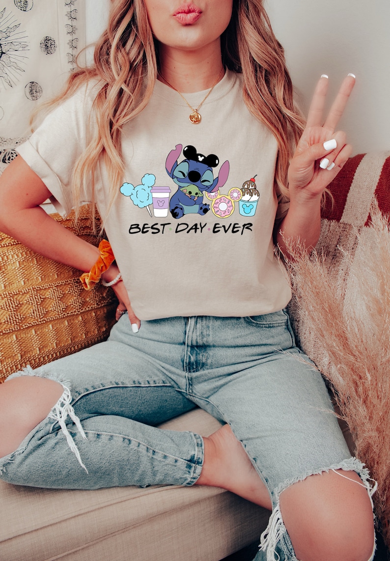 K&ouml;nnte beinhalten: Eine Frau tr&auml;gt ein hellbeiges T-Shirt mit einer blauen Comicfigur, die einen gr&uuml;nen Au&szlig;erirdischen h&auml;lt und von Comicbildern von Essen und Getr&auml;nken umgeben ist. Der Text "BEST DAY EVER" ist unter der Comicfigur gedruckt.