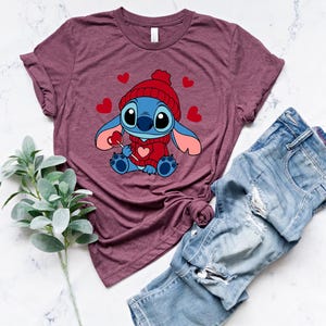 May include: Burgundy t-shirt featuring a cartoon character wearing a red hat and sweater, holding a heart-shaped lollipop. The design includes red hearts. Light blue denim jeans are next to the shirt. A sprig of green leaves is also in the image.