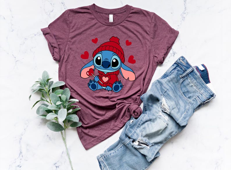 May include: Burgundy t-shirt featuring a cartoon character wearing a red hat and sweater, holding a heart-shaped lollipop. The design includes red hearts. Light blue denim jeans are next to the shirt. A sprig of green leaves is also in the image.