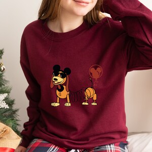 May include: A maroon sweatshirt with a cartoon image of a dog wearing sunglasses and a Mickey Mouse balloon. The dog is a spring-loaded toy with a coiled spring tail.