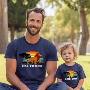 May include: A father and son wearing matching navy blue t-shirts with a retro sunset graphic. The father's shirt says "Like Father" and features a lion. The son's shirt says "Like Son" and features a smaller lion.