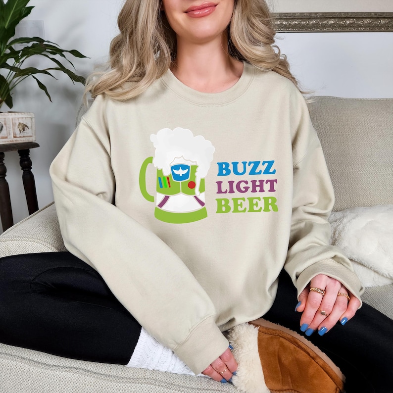 May include: A light beige sweatshirt featuring a graphic of a green beer mug with a cartoon character inside. The text "Buzz Light Beer" is printed in colourful letters on the sweatshirt.