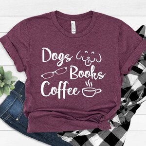May include: A maroon short-sleeved t-shirt featuring white screen-printed text that reads Dogs, Books, and Coffee. The design includes a line-art graphic of a dog face, a pair of eyeglasses, and a steaming coffee cup centered on the chest.