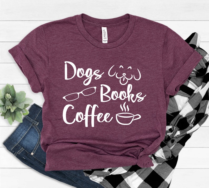 May include: A maroon short-sleeved t-shirt featuring white screen-printed text that reads Dogs, Books, and Coffee. The design includes a line-art graphic of a dog face, a pair of eyeglasses, and a steaming coffee cup centered on the chest.