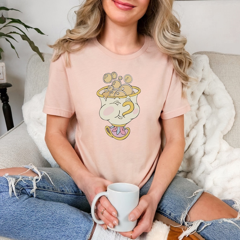Puede incluir: Una camiseta rosa con una imagen de dibujos animados de una taza de t&eacute; con una cara y burbujas que salen de ella. La taza de t&eacute; tiene un borde dorado y un asa rosa y blanca.