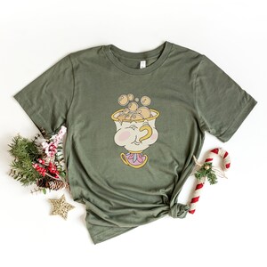 Puede incluir: Una camiseta verde con una imagen de dibujos animados de Chip de La Bella y la Bestia. Chip sostiene una taza de t&eacute; con burbujas que salen de ella. La camiseta est&aacute; anudada.