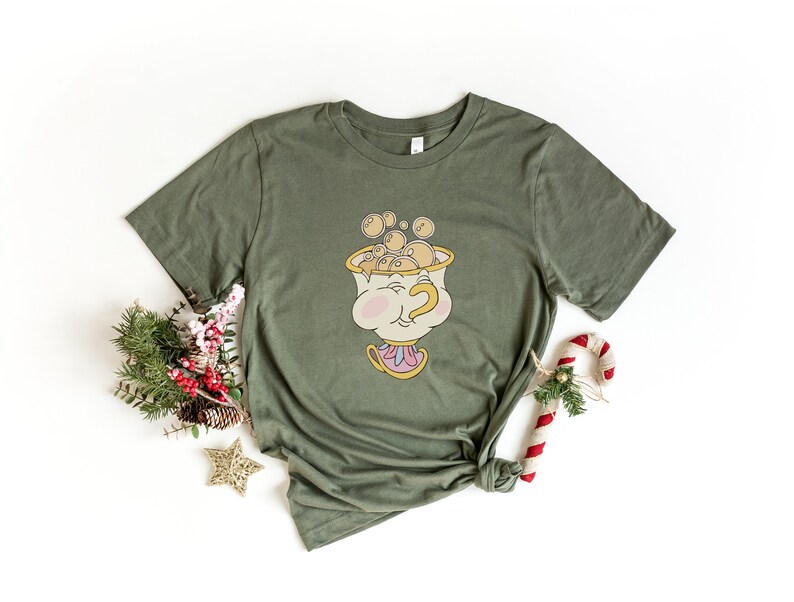 Puede incluir: Una camiseta verde con una imagen de dibujos animados de Chip de La Bella y la Bestia. Chip sostiene una taza de t&eacute; con burbujas que salen de ella. La camiseta est&aacute; anudada.